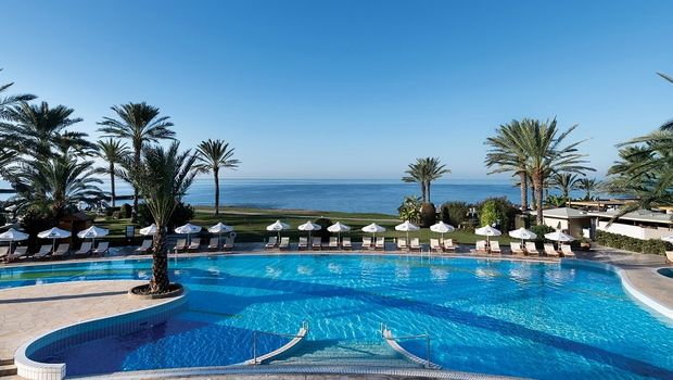 Großer Pool mit Palmen und Sonnenliegen, dahinter eine weitläufige Rasenfläche und der Blick auf das ruhige Meer.