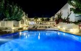 Stimmungsvoll beleuchteter Hotelpool bei Nacht – weiße Gebäude im Kykladenstil, umgeben von Palmen, Bougainvillea und gemütlichen Liegen.