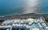 Luftaufnahme des Costa Grand Resort & Spa auf Santorin – elegante Hotelanlage direkt am schwarzen Lavastrand von Kamari mit mehreren Pools, weißen Gebäuden im Kykladenstil und herrlichem Blick auf das funkelnde Meer.
