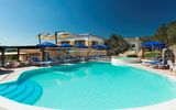 Einladender Außenpool mit türkisblauem Wasser, umgeben von blauen Sonnenschirmen und Liegestühlen vor einer mediterranen Hotelanlage unter klarem Himmel.