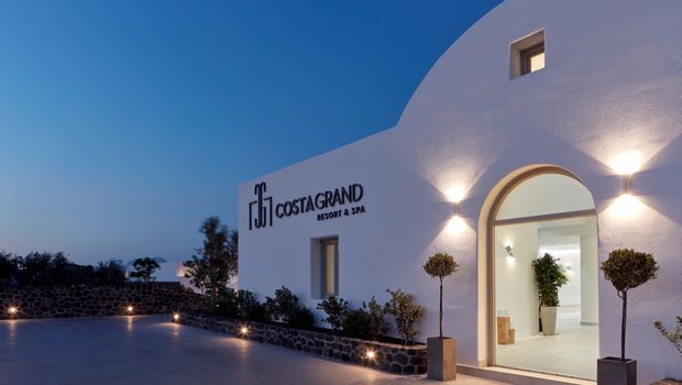 Beleuchteter Hoteleingang des Costa Grand Resort & Spa am Abend – modernes, weißes Gebäude im Kykladenstil mit stilvoller Außenbeleuchtung und mediterraner Bepflanzung.