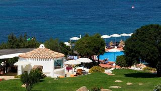 Blick auf einen idyllischen Hotelgarten mit mediterranen Pflanzen, Poolbereich mit Sonnenliegen und weißen Sonnenschirmen – im Hintergrund das tiefblaue Meer, das zum Träumen einlädt.