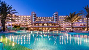 Beleuchtetes Hotel mit großem Poolbereich bei Nacht, eingerahmt von Palmen und klarer Abendstimmung.