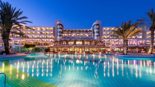 Beleuchtetes Hotel mit großem Poolbereich bei Nacht, eingerahmt von Palmen und klarer Abendstimmung.