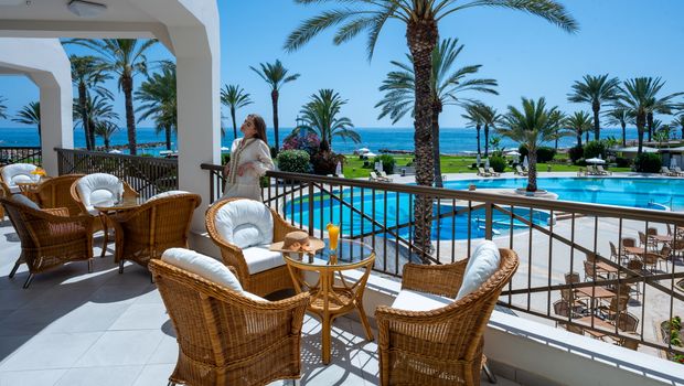 Terrasse mit gemütlichen Rattanmöbeln und Meerblick – im Hintergrund der Poolbereich, Palmen und das glitzernde Mittelmeer.