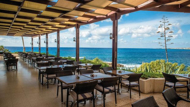 Überdachte Außenterrasse mit dunklen Holztischen und herrlichem Blick auf das tiefblaue Meer.