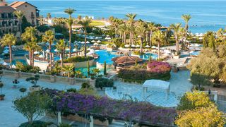 Großzügige Hotelanlage mit mehreren Pools, Palmen, weißen Sonnenschirmen und Meerblick – eine mediterrane Oase zum Entspannen.