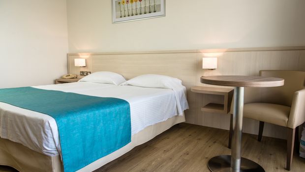 Helles Doppelzimmer mit Holzboden, schlichter Einrichtung und türkisfarbenem Bettläufer.